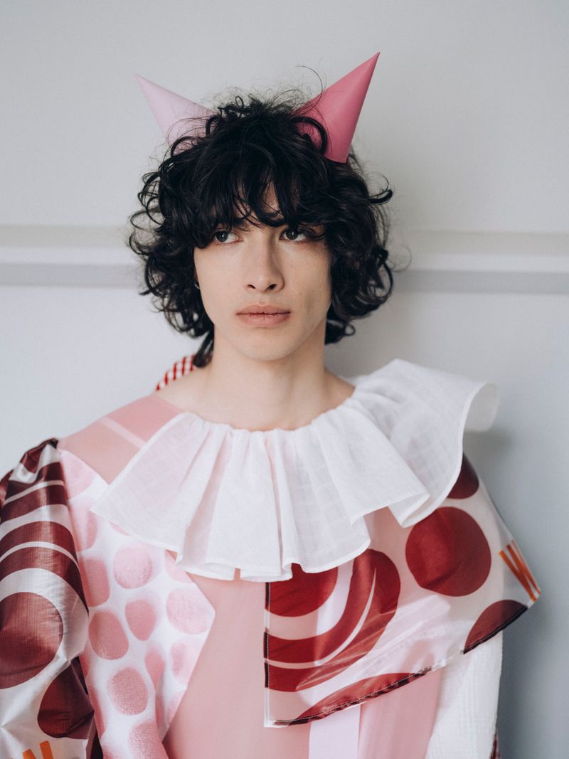 A young adult poses indoors wearing a pink costume with white ruff collar and pink cone hat. Soft lighting, portrait-style framing.
