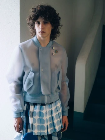 Fashionable young adult man poses indoors wearing a light blue, translucent jacket and patterned skirt, looking directly at the camera.