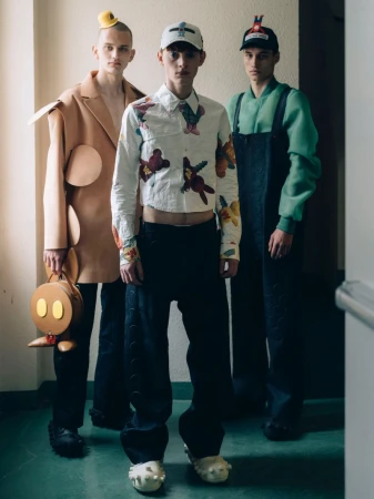 Three young men pose indoors in eclectic streetwear outfits with hats, patterned shirts, black pants and shoes, against a white wall.