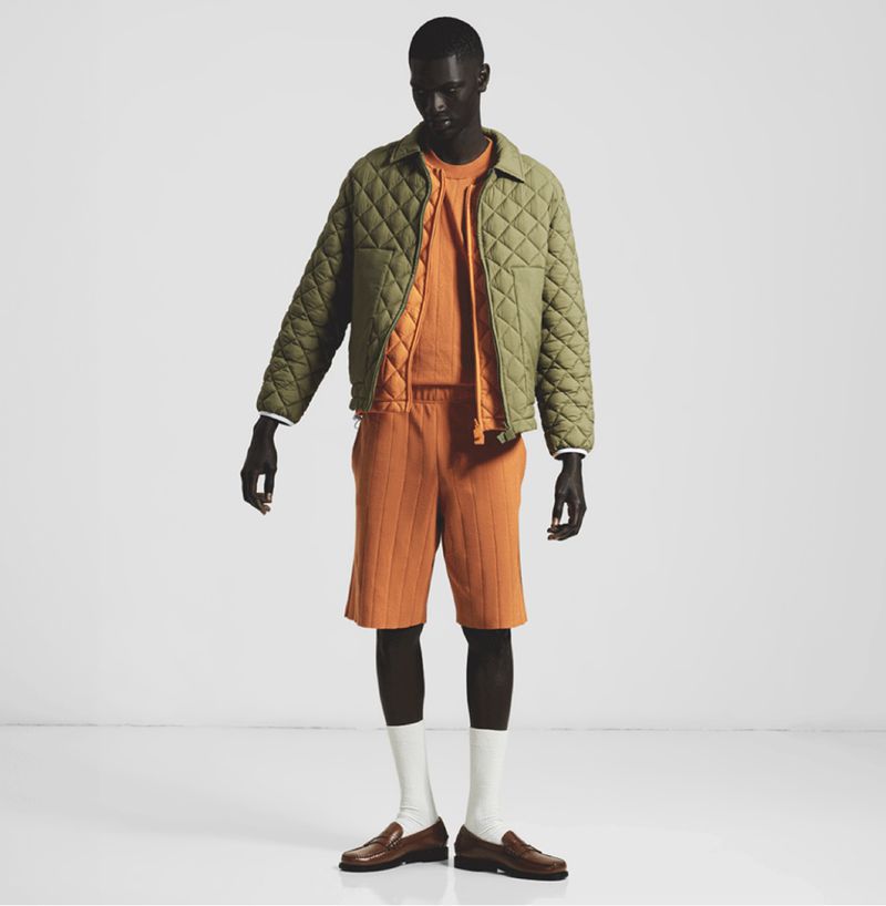 Dark-skinned male model poses in a sage quilted jacket, light-orange ribbed knitwear, shorts, white socks, and brown loafers on a white backdrop.