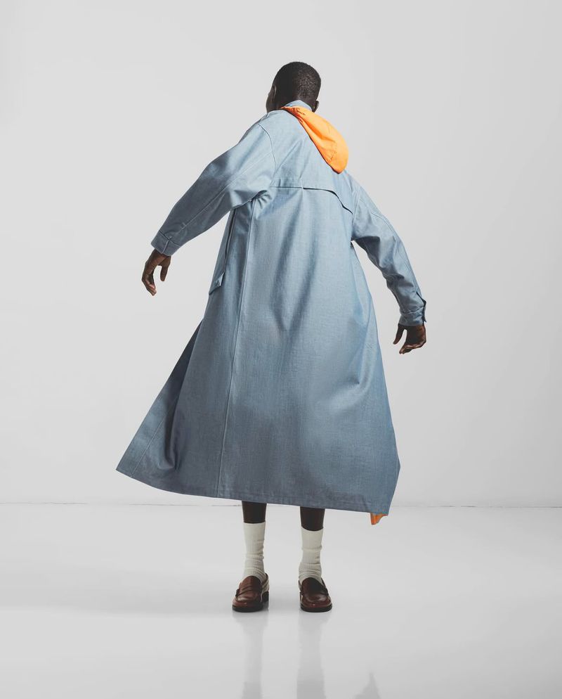 An adult male model, from the rear, wears a long blue denim coat with a bright orange hood, with dark trousers, white socks, and brown leather shoes.