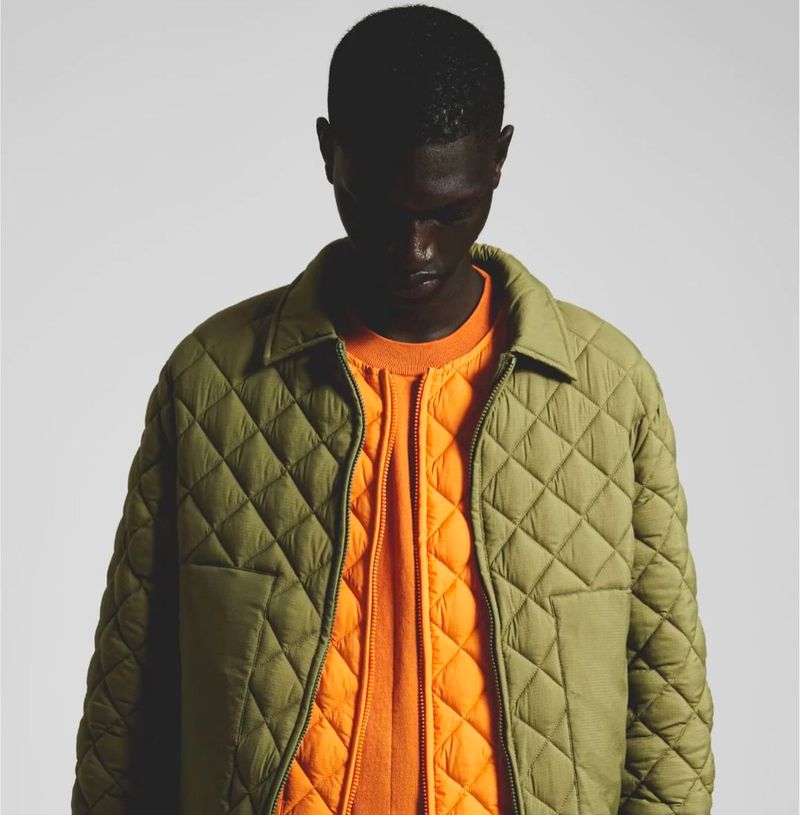 An adult male models an olive green quilted jacket over an orange quilted crewneck sweater in a studio with a neutral backdrop.
