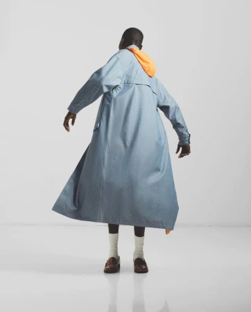 Model seen from behind wearing a long blue coat with an orange hood, white socks, and brown loafers against a plain white background.