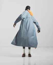 Model seen from behind wearing a long blue coat with an orange hood, white socks, and brown loafers against a plain white background.
