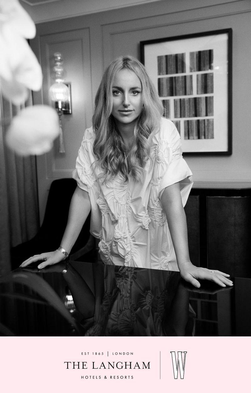 Black and white portrait of a woman with long blonde hair leaning on a shiny black table in front of a framed art piece.