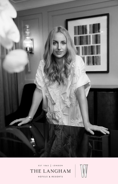 Black and white portrait of a woman with long blonde hair leaning on a shiny black table in front of a framed art piece.