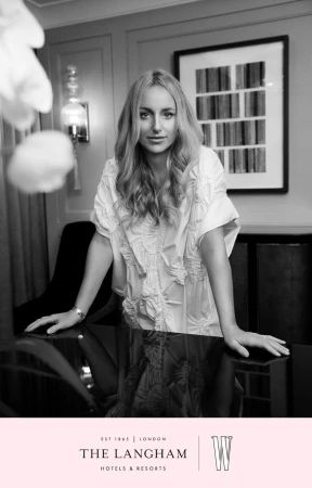 Black and white portrait of a woman with long blonde hair leaning on a shiny black table in front of a framed art piece.