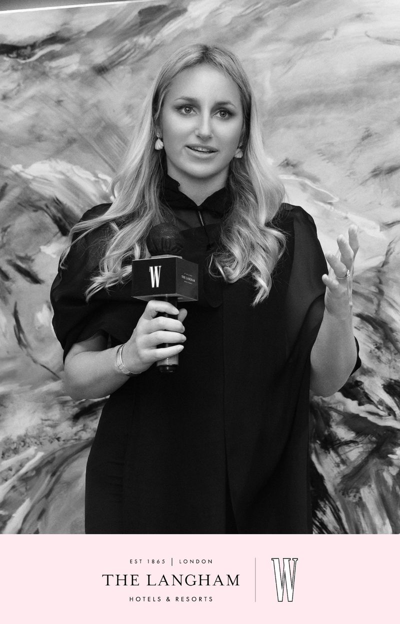 Portrait of a blonde woman in a black blouse speaking into a branded microphone in front of an abstract monochrome backdrop.