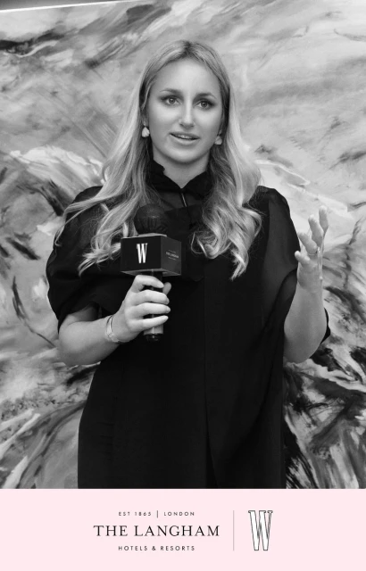 Portrait of a blonde woman in a black blouse speaking into a branded microphone in front of an abstract monochrome backdrop.
