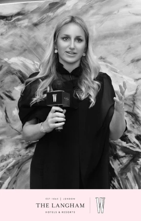 Portrait of a blonde woman in a black blouse speaking into a branded microphone in front of an abstract monochrome backdrop.