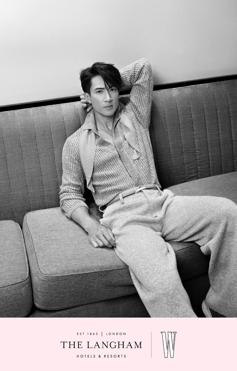 Black and white studio portrait of an adult male reclining on a gray couch wearing a knitted sweater, pants, and a belt.