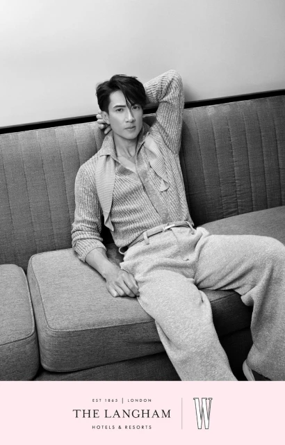Black and white studio portrait of an adult male reclining on a gray couch wearing a knitted sweater, pants, and a belt.
