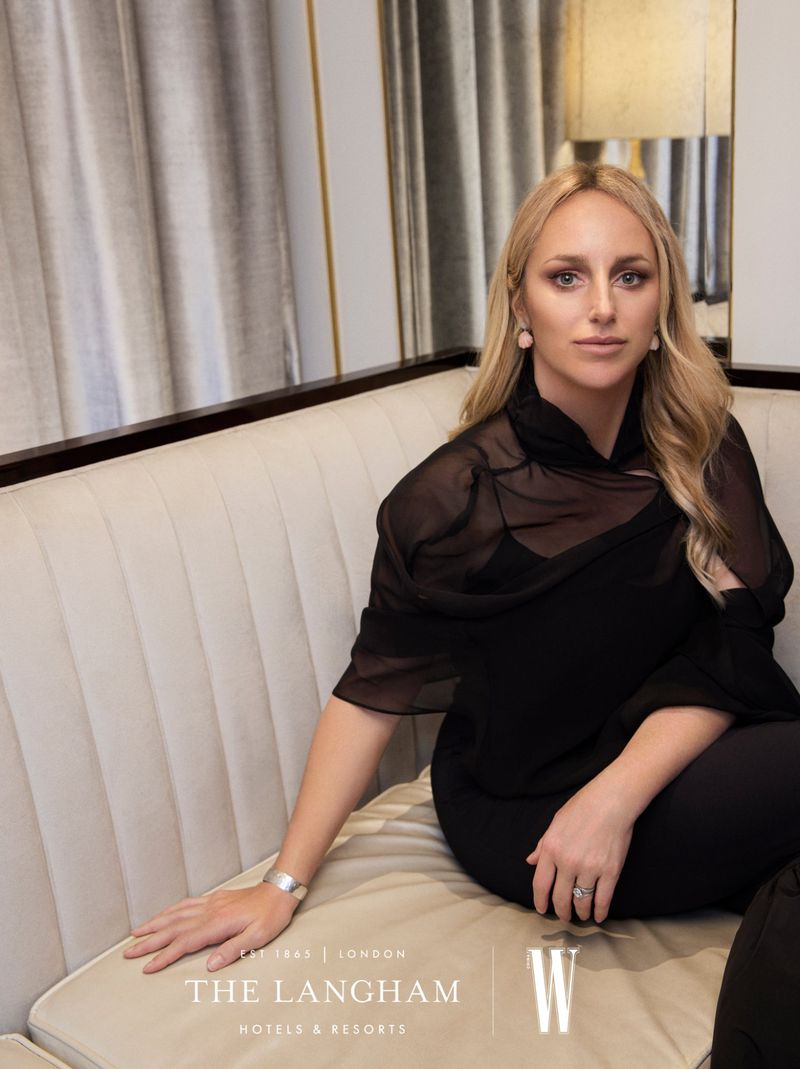 Blonde woman in black outfit sits on a cream sofa, posing indoors with neutral decor and soft, diffused lighting in a hotel setting.