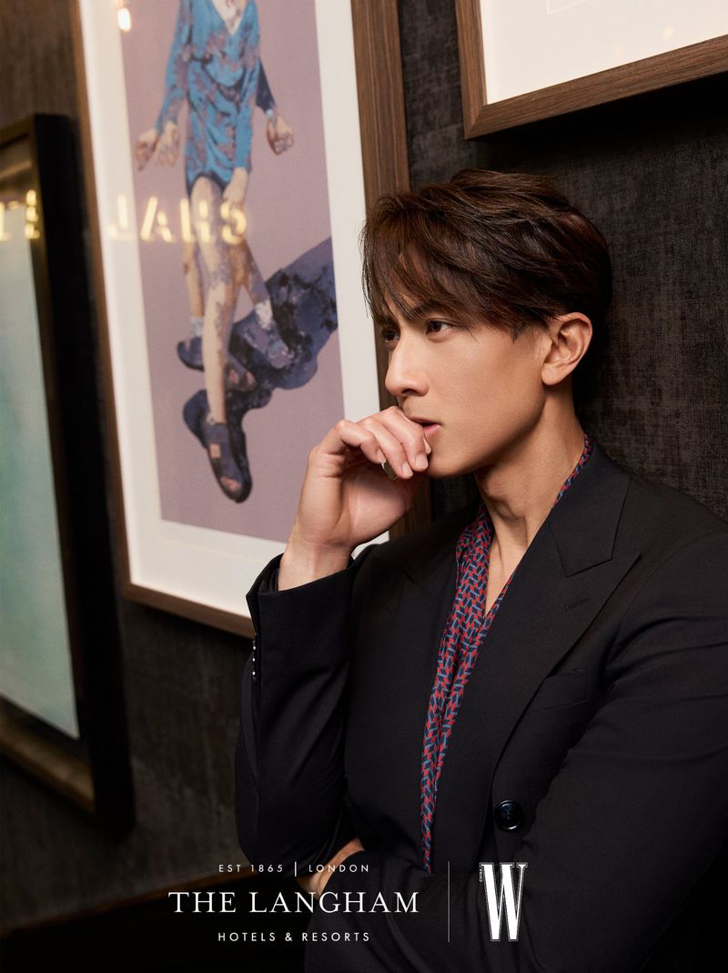 Portrait of a young man in a suit jacket against a patterned wall with framed artwork inside of The Langham Hotel in London.