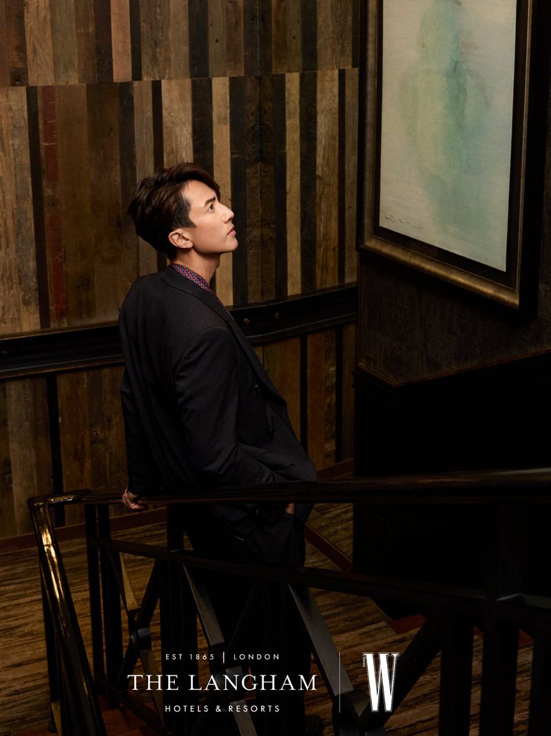 Adult male in suit jacket looks at framed painting on wall inside The Langham Hotel in London with patterned wood wall and banister.