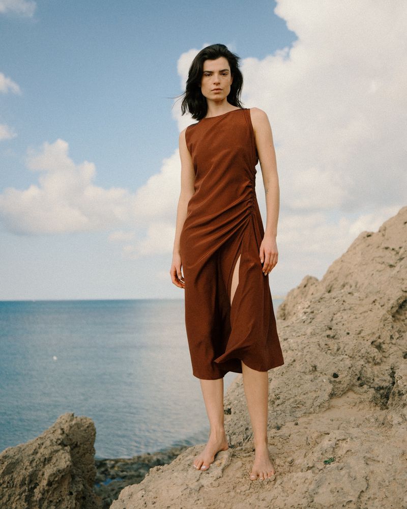 A woman stands on a rocky cliff in a brown dress with a thigh slit, posing against a cloudy sky and ocean backdrop.