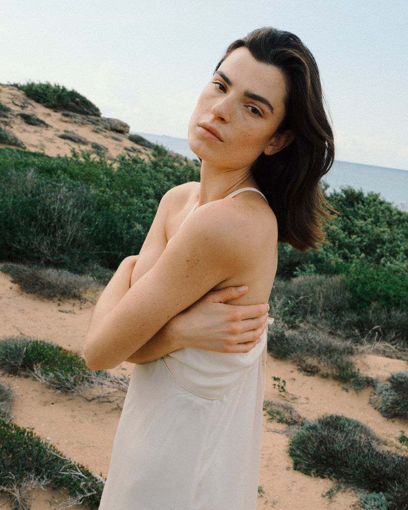 Woman with dark hair wearing a cream slip dress with arms crossed against a coastal dunes background.