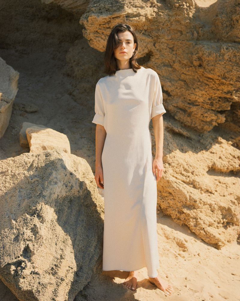 A slender woman with dark hair, wearing a cream-colored midi dress, stands barefoot among tan rocks, illuminated by natural lighting.