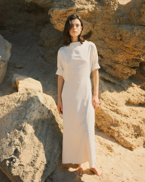 A slender woman with dark hair, wearing a cream-colored midi dress, stands barefoot among tan rocks, illuminated by natural lighting.
