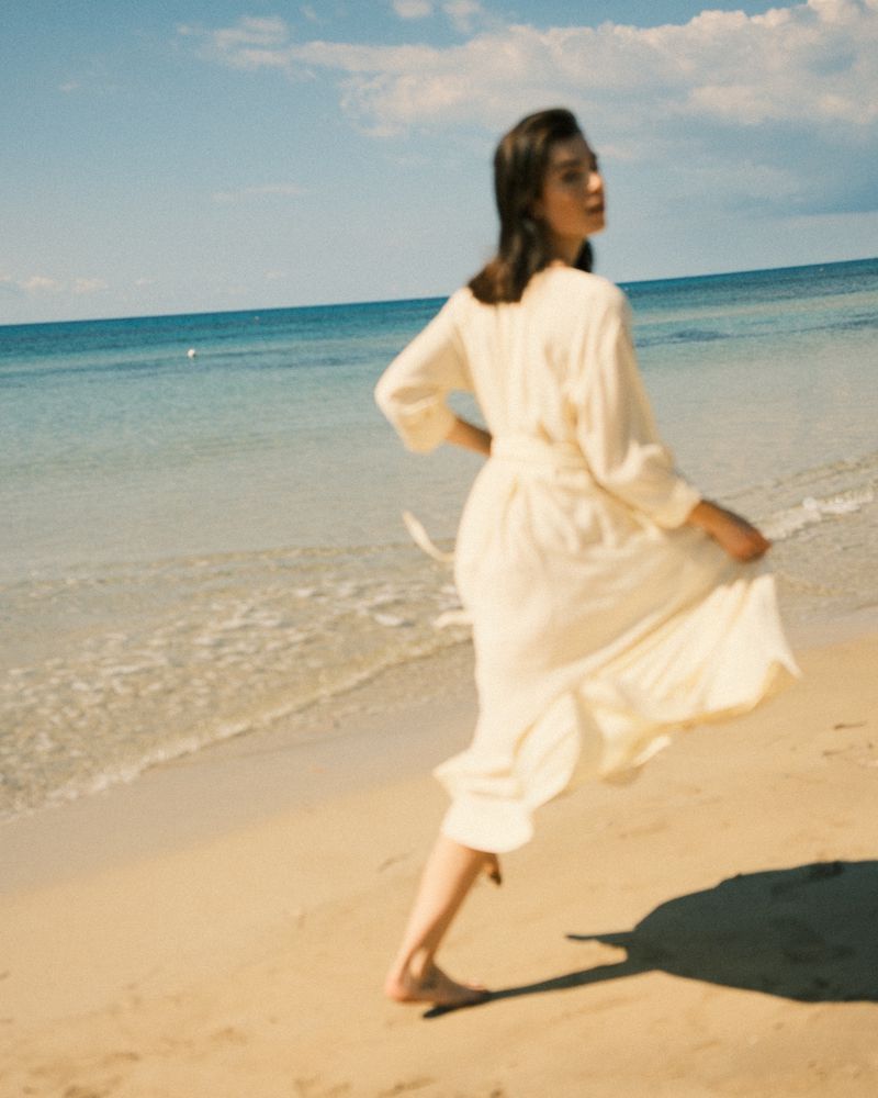 Model in a cream wrap dress walking on a sandy beach by the ocean under a clear sky.