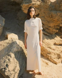Barefoot woman wearing an off-white high-neck maxi dress standing on sand against sunlit sandstone rocks.