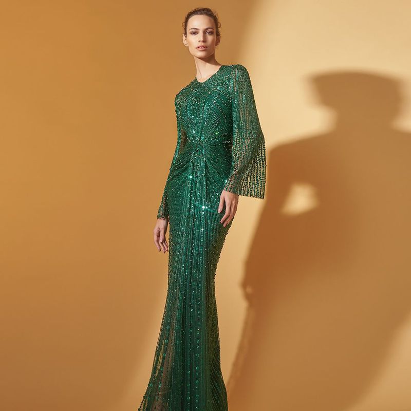Elegant woman in a shimmering emerald green gown with flowing sleeves, standing against a soft tan background, fashion and style.