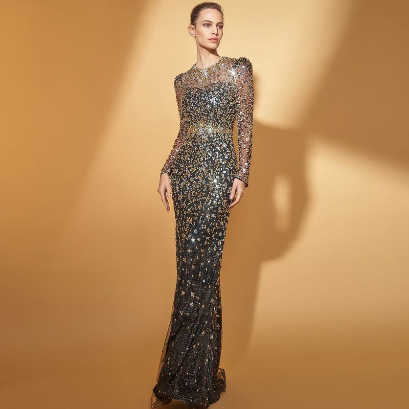 A slender woman poses in a shimmering black and gold sequined gown against a warm beige backdrop in a fashion editorial shot.