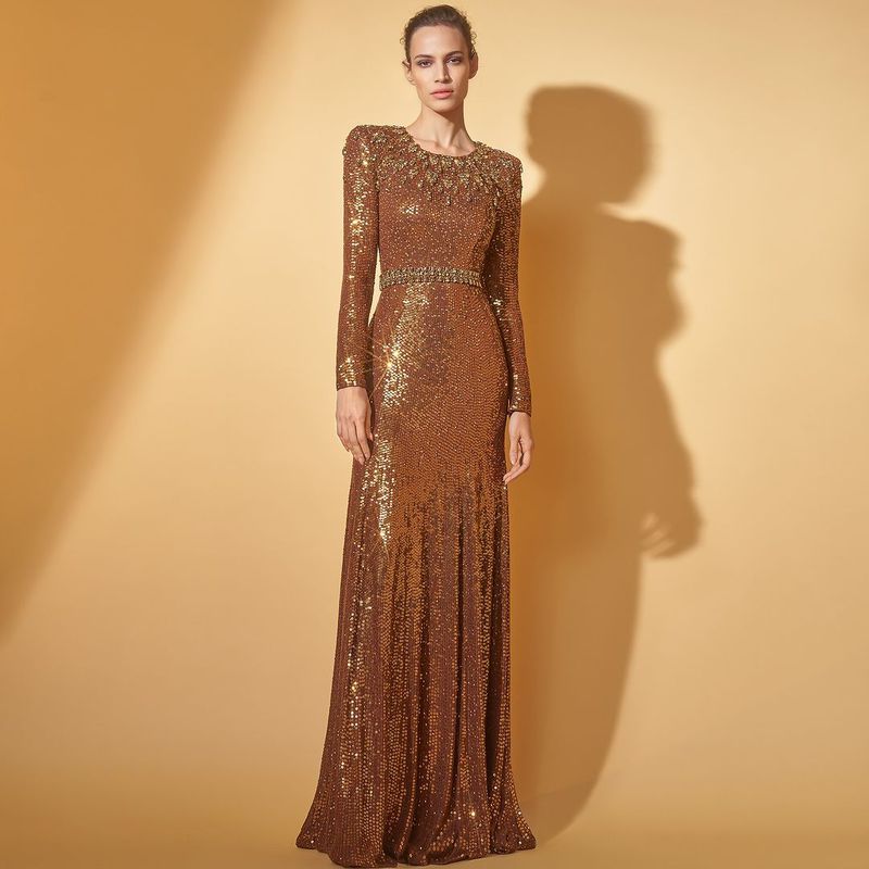 Elegant adult woman in a shimmering bronze-gold, long-sleeve gown with jewel-encrusted collar and belt, standing in a neutral studio setting.