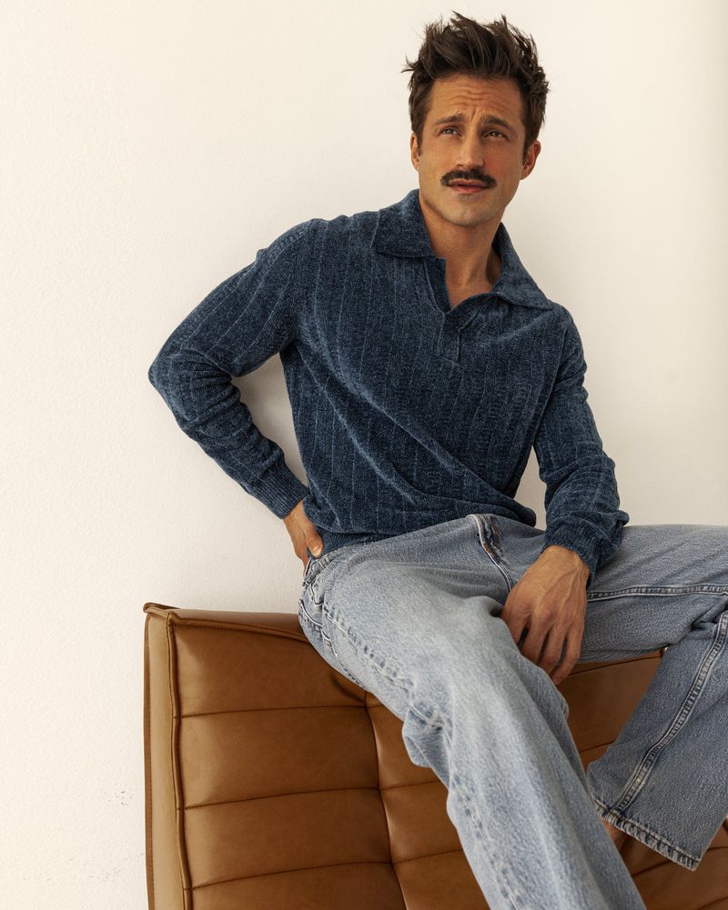 Adult man with a mustache poses indoors, seated on a brown leather couch, wearing a blue sweater and jeans against a white wall backdrop.
