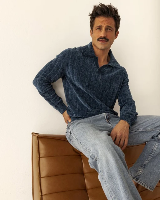 Adult man with a mustache poses indoors, seated on a brown leather couch, wearing a blue sweater and jeans against a white wall backdrop.