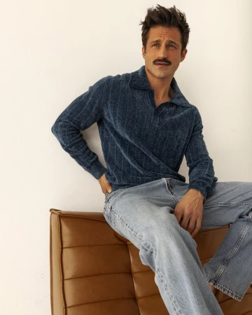 Adult man with a mustache poses indoors, seated on a brown leather couch, wearing a blue sweater and jeans against a white wall backdrop.