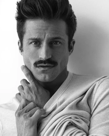 Man with styled dark hair and a mustache poses wearing a waffle-knit shirt in a black-and-white studio portrait.