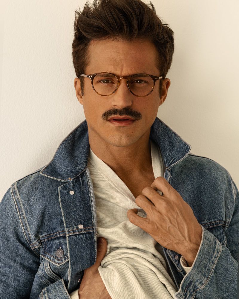Portrait of a man wearing glasses and a denim jacket, posing indoors against a neutral wall, showcasing a casual yet stylish aesthetic.