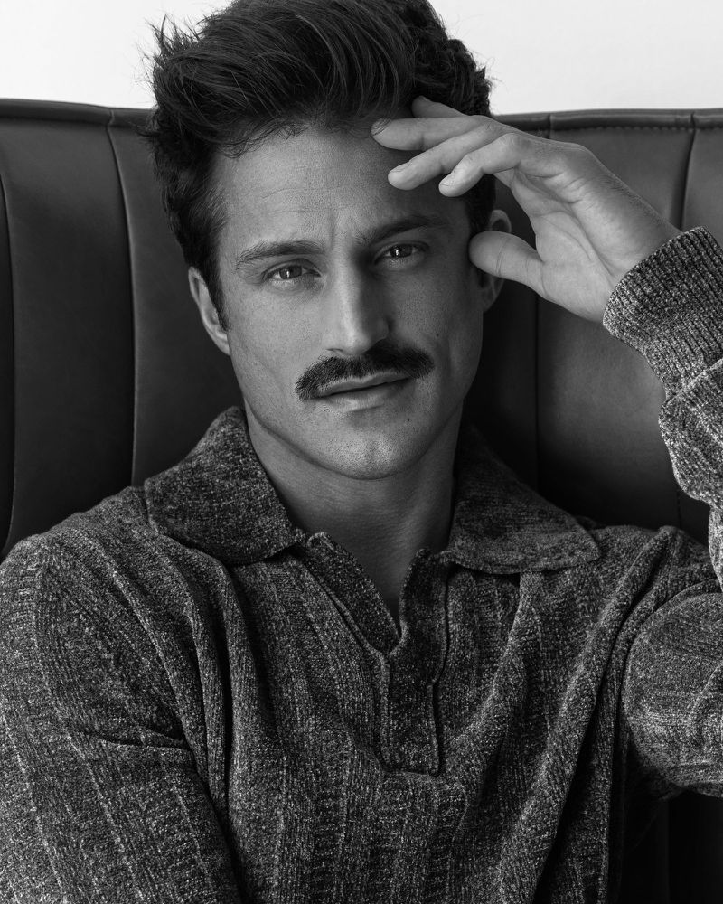 Monochrome close-up portrait of a man with a mustache wearing a textured knit polo shirt resting his hand against his forehead.