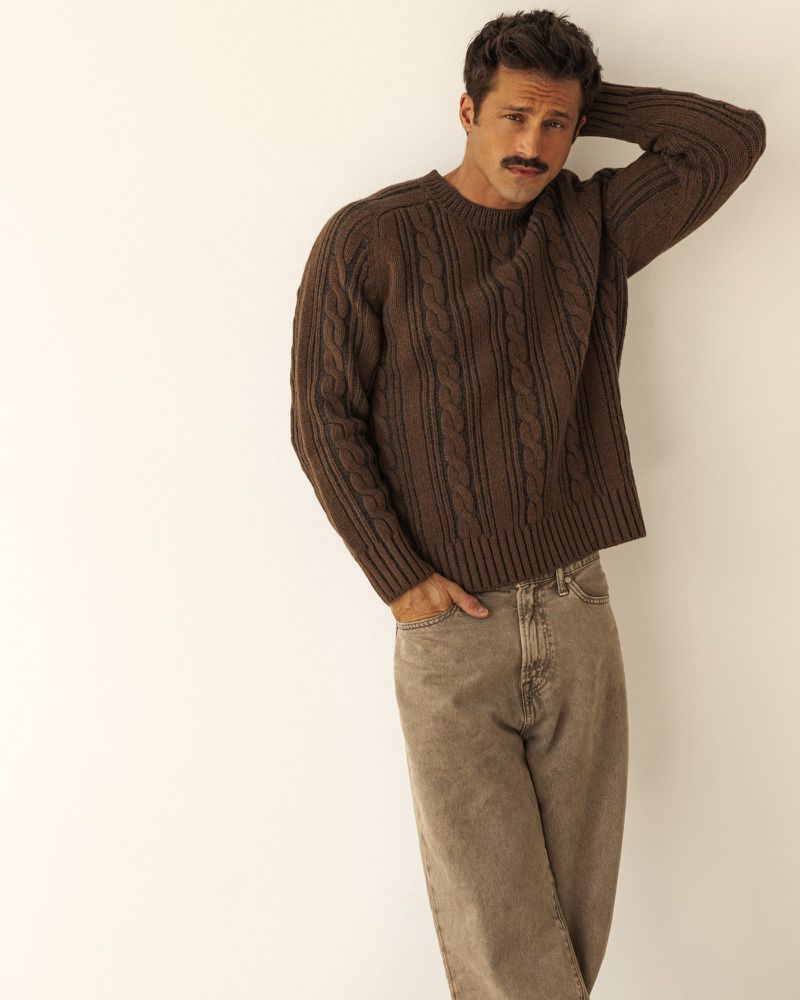 Man with a mustache wearing a dark brown cable knit sweater and beige jeans posing against an off-white studio background.