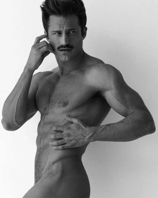 Black-and-white studio shot of a muscular adult male with a mustache, posing shirtless with hand raised to head and hand on ribs.