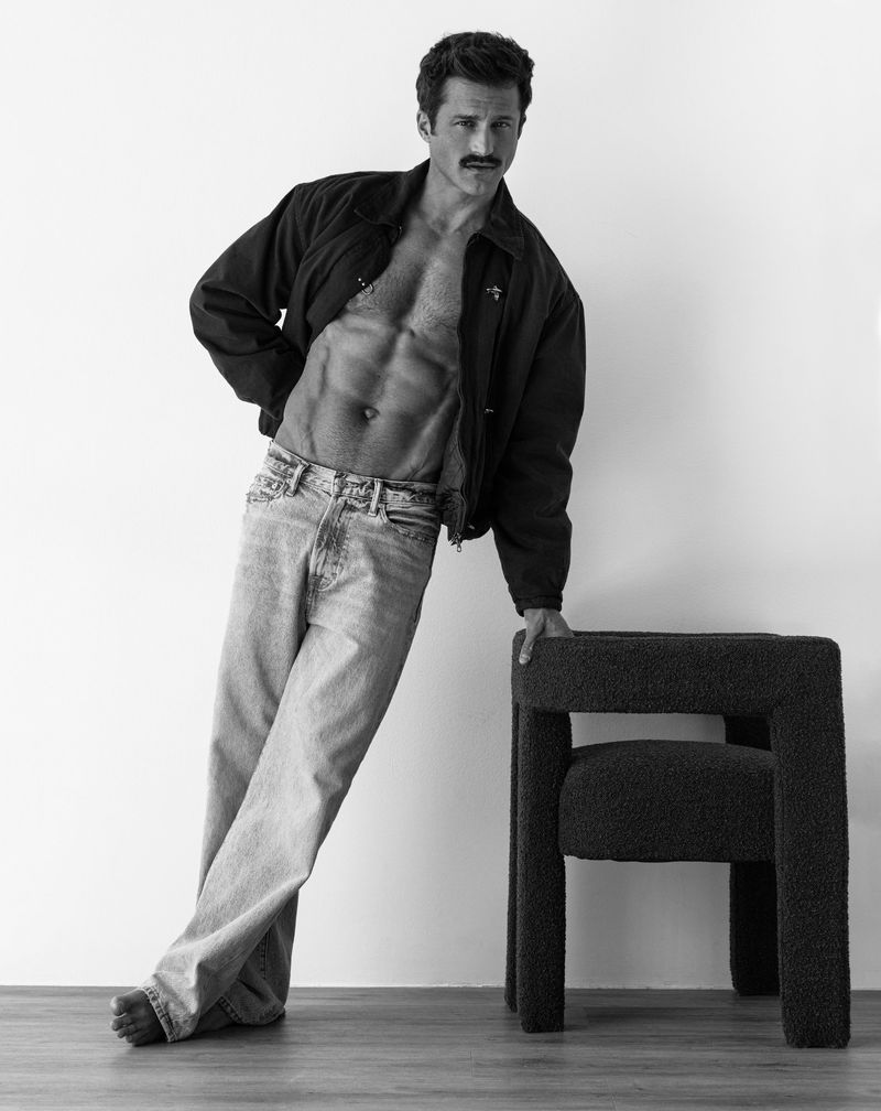 Monochrome portrait of a shirtless man with a mustache wearing a dark open jacket and light jeans leaning on a dark stool