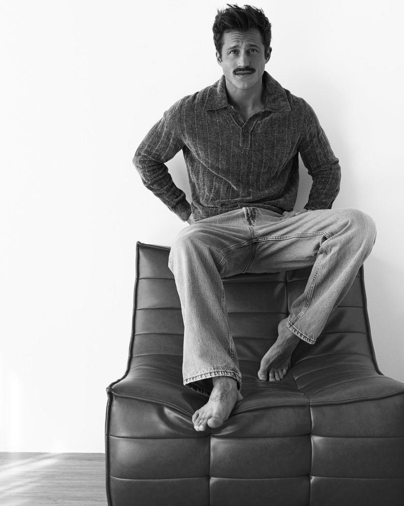 Black and white portrait of a man with a mustache sitting on a black leather chair in a studio, wearing a sweater and jeans.