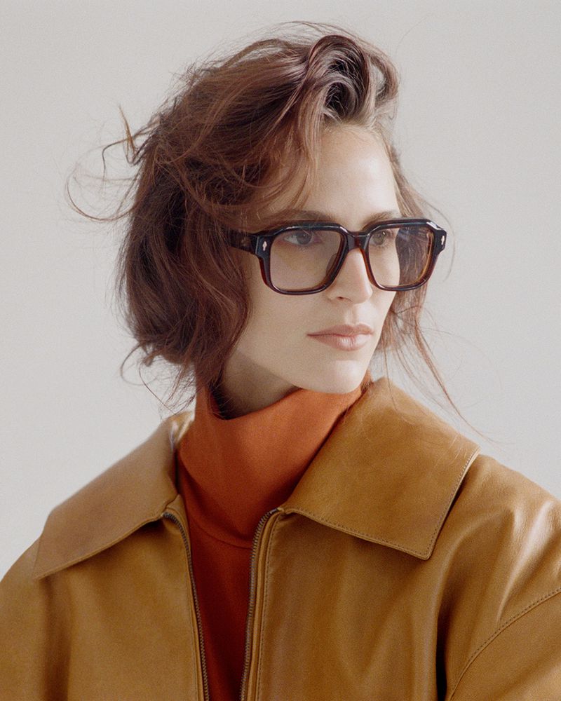 A fashionable adult female with medium auburn hair is wearing square-framed glasses, a turtleneck, and a light brown leather jacket.