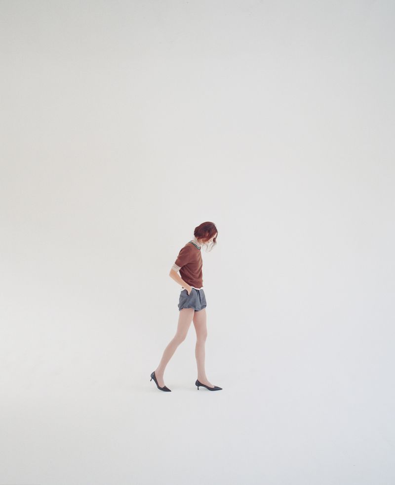 A woman with auburn hair walks across a light gray seamless backdrop, wearing a brown t-shirt, blue shorts, and black heels.