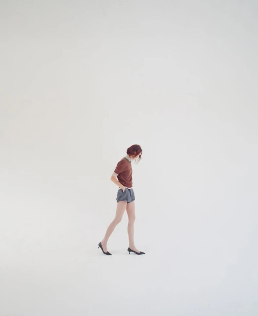 A woman with auburn hair walks across a light gray seamless backdrop, wearing a brown t-shirt, blue shorts, and black heels.