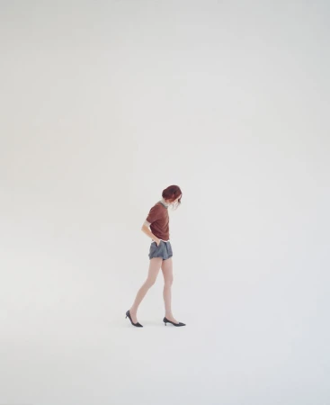 A woman with auburn hair walks across a light gray seamless backdrop, wearing a brown t-shirt, blue shorts, and black heels.