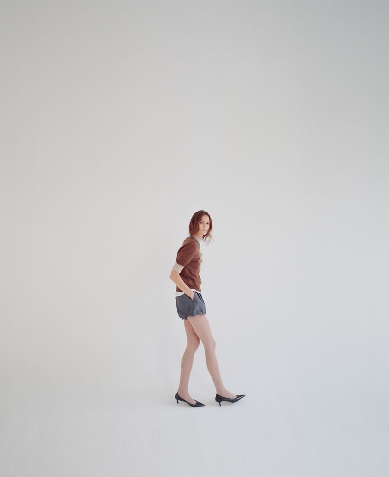 A young adult woman posing in a brown top and blue shorts, standing in front of a plain white background in a studio setting.