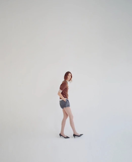 A young adult woman posing in a brown top and blue shorts, standing in front of a plain white background in a studio setting.
