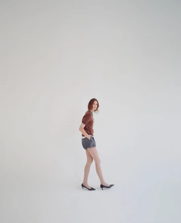 A young adult woman posing in a brown top and blue shorts, standing in front of a plain white background in a studio setting.