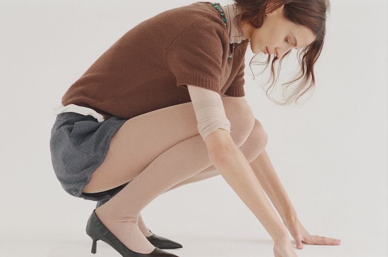 Woman in brown sweater, grey shorts and black heels squats in photo studio against a white backdrop, looking down in a fashion pose.