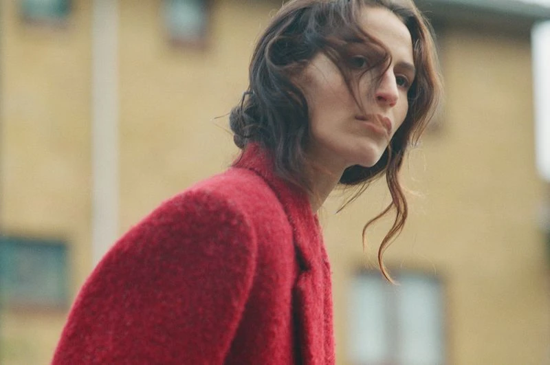 A woman with dark hair and a red coat is captured in a medium shot, against a blurred background of a yellow building and sky.