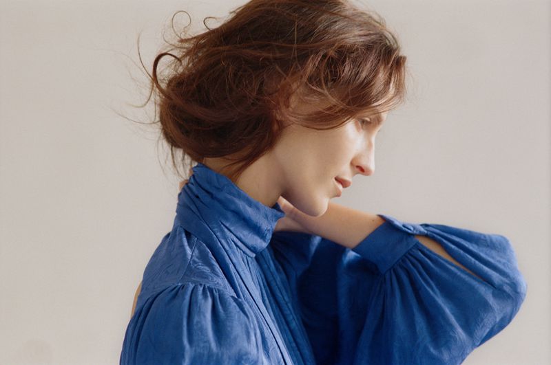 Profile view of a woman with auburn hair in a blue high-neck blouse, her hand gently touching her neck in a light, airy indoor setting.