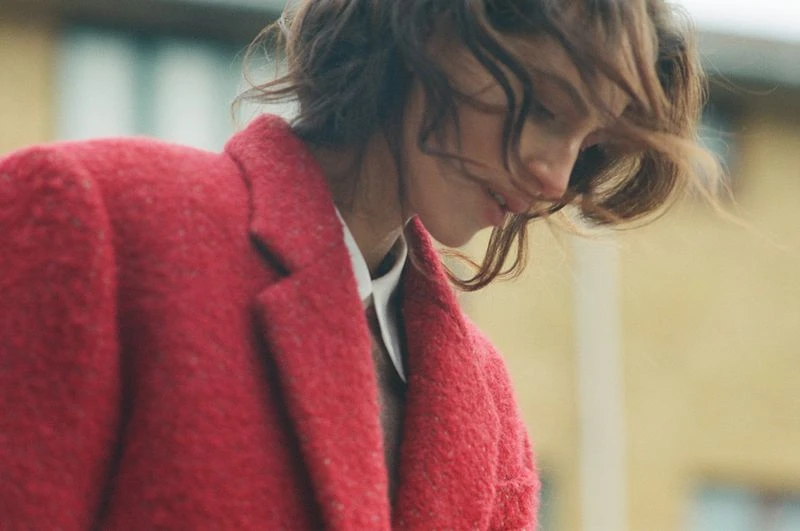 Woman in a red coat posing outside. The woman is looking down and to the right, with windblown hair. Medium shot with blurred background.