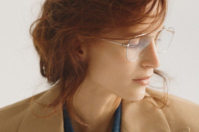 Close-up of a stylish adult woman with red hair and glasses wearing a jacket in a bright indoor setting. Beauty shot.